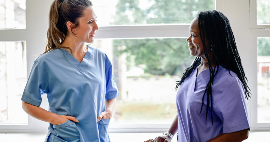 Two nurses are standing next to each other in front of a window and talking