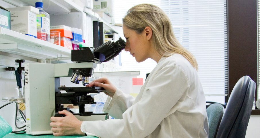  A woman is sitting at a desk in a medical lab looking through a microscope