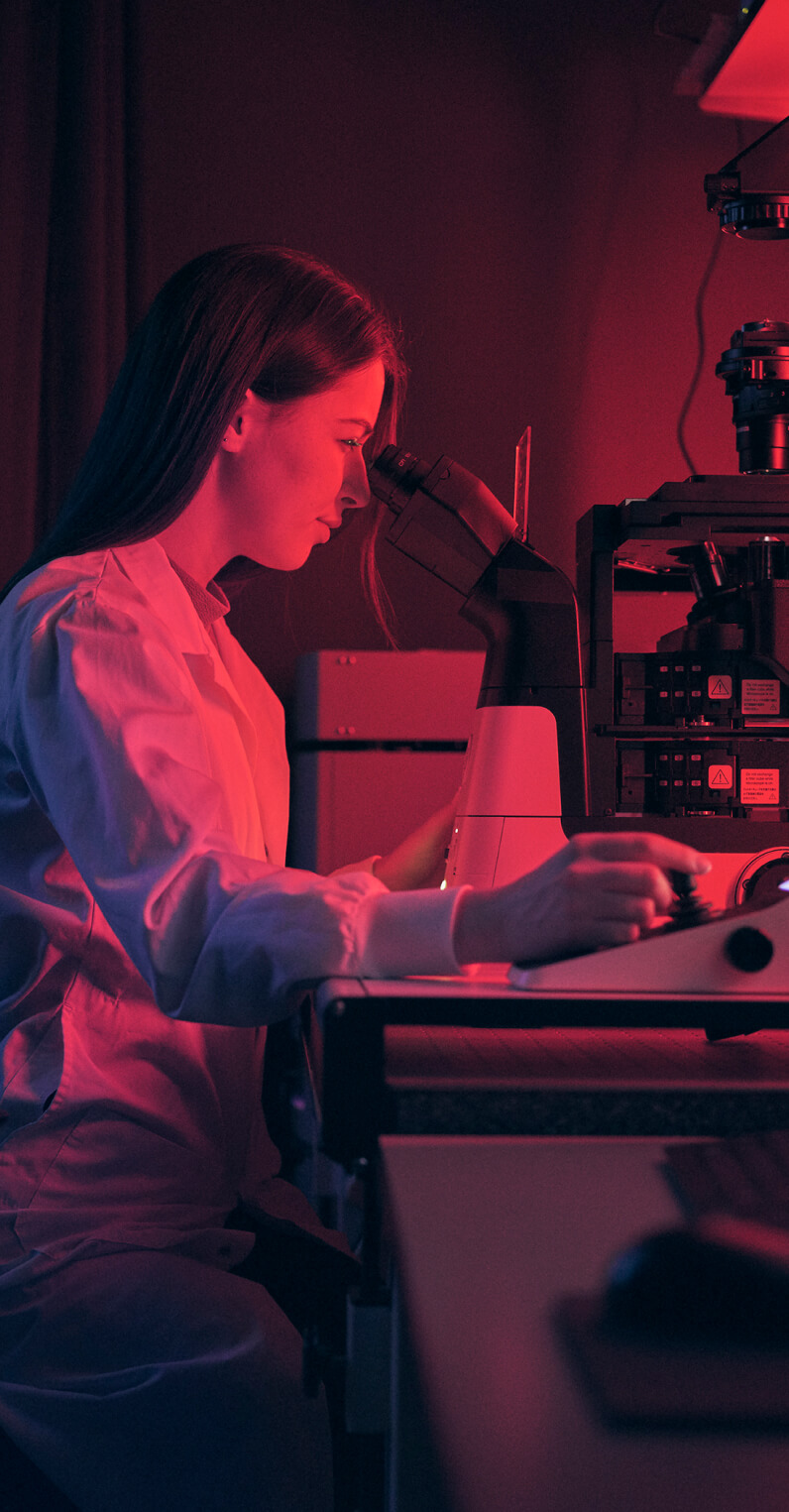 Person in a lab coat working with a microscope under red lighting.